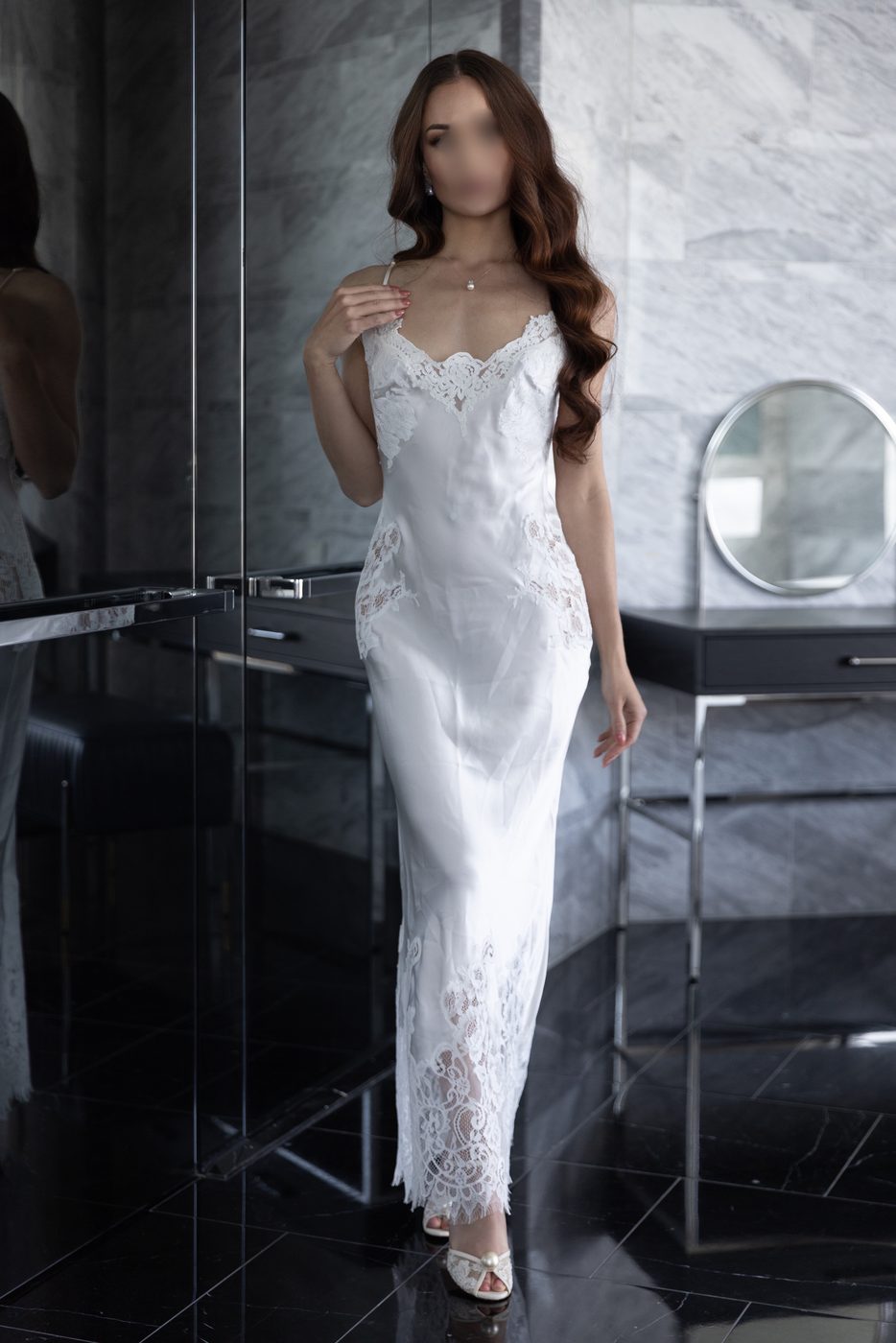 Irina in a white lace-trimmed silk slip gown and pearl jewellery, standing beside a round mirror in a marble bathroom
