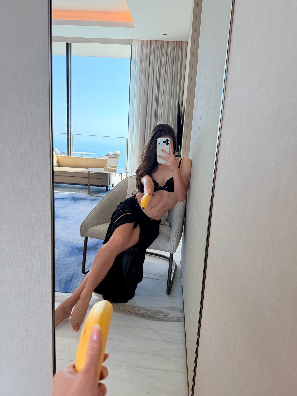 Irina, a seated mirror selfie holding a yellow phone in a bright coastal suite